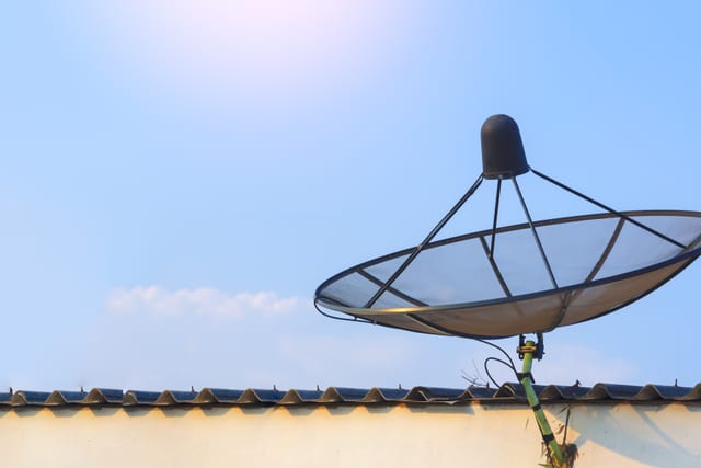 Black mesh satellite dish mounted above a tiled roofline against a pale blue sky