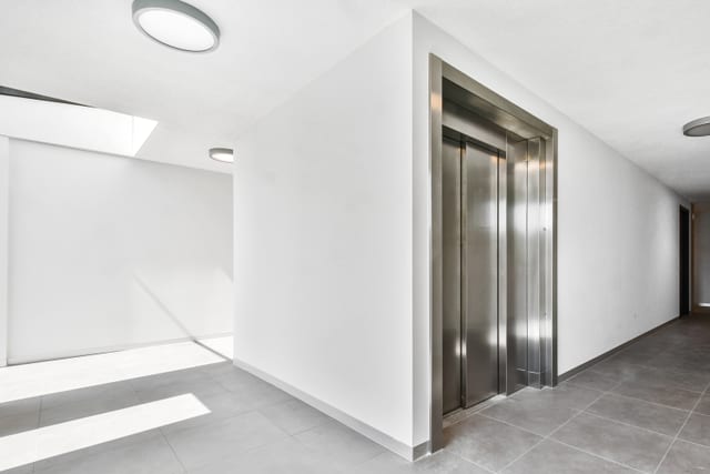 Brushed stainless lift doors recessed into a white wall on an empty apartment landing, with grey floor tiles and daylight from a skylight