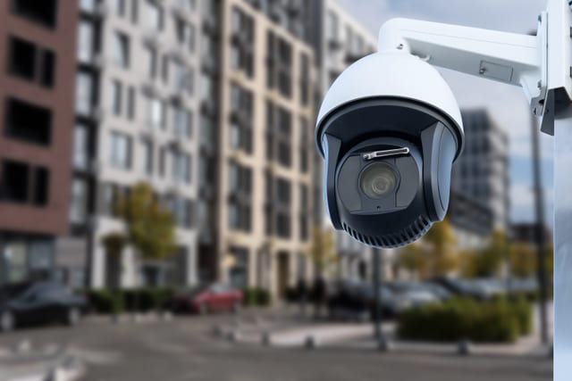 White dome security camera on a bracket, with balconied apartment blocks and street trees out of focus behind it