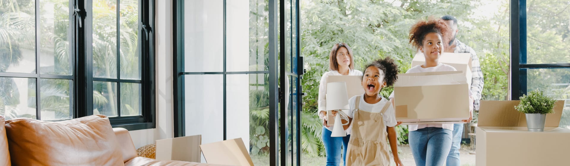 A child in tan overalls walks through an open glass door into a living room while an older child and two adults follow carrying cardboard boxes and a table lamp, with a tan leather sofa at the left and stacked boxes topped by a small potted plant at the right.