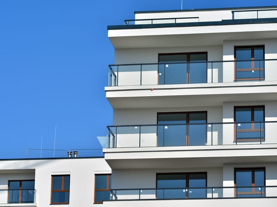 Upper floors of a modern white apartment block, its stepped balconies fronted with clear glass balustrades and dark grey handrails, warm wood-framed windows catching the sun against a deep blue sky.