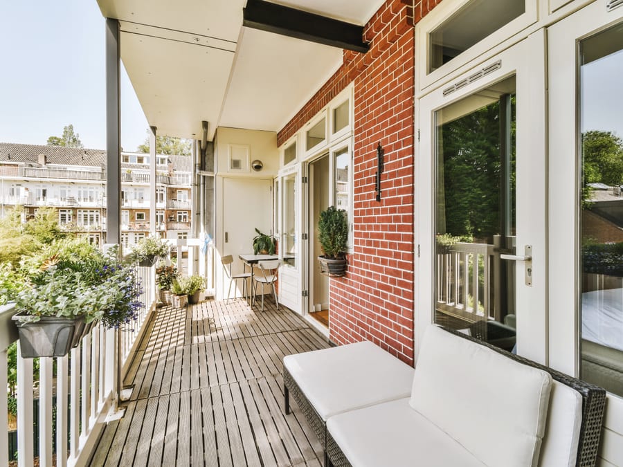 A covered apartment balcony with a white painted railing holding planter boxes of flowers, a slatted timber deck, a small bistro table and two chairs, and a facing row of brick buildings with white balconies across a green courtyard.