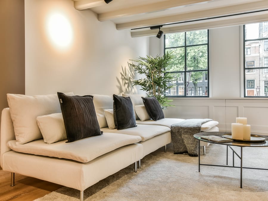 A sunlit apartment living room with a low cream sectional sofa, dark charcoal cushions, a round glass-topped coffee table holding pillar candles on a brass tray, and tall black-framed windows onto a leafy street.