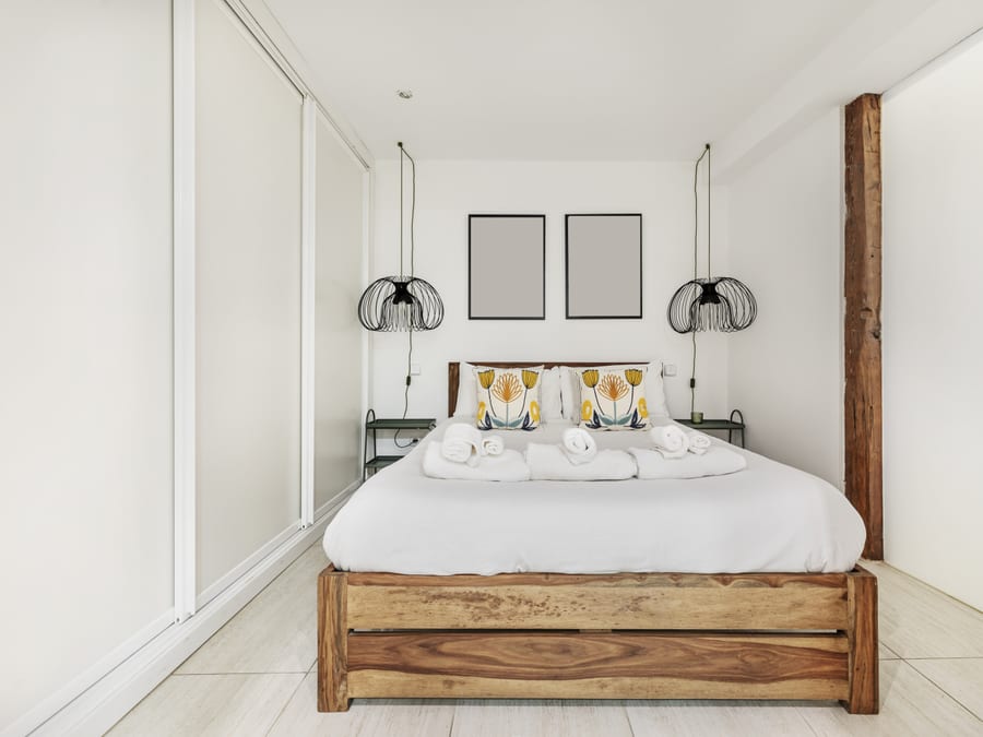 A compact white studio with a solid-wood bed frame and crisp white bedding, two black wire cage pendants hanging either side and a pair of framed prints centred above the headboard.