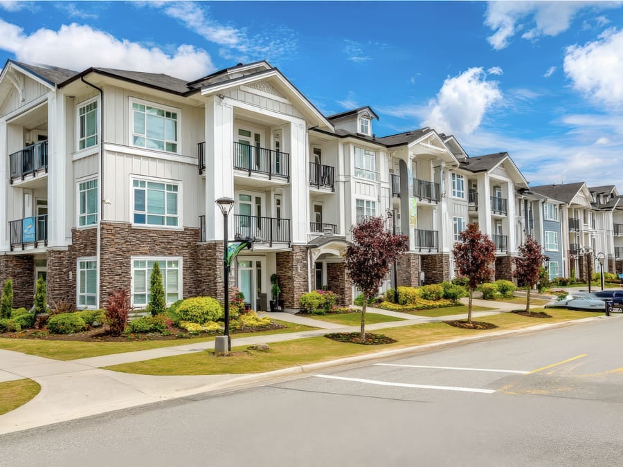 A long three-storey apartment community in white board-and-batten siding with stacked stone bases, black balcony railings and gabled porches, running down a landscaped street of clipped shrubs and lamp posts.