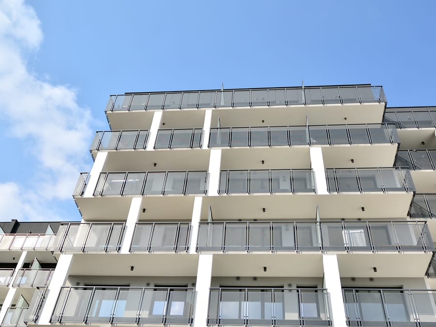 A cream and white multi-storey apartment building photographed from below, showing five levels of balconies with clear glass railings and white piers between them against a blue sky.