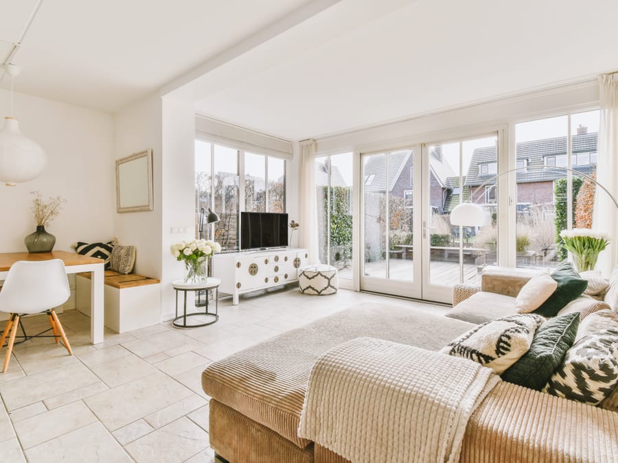 A bright open-plan living and dining room with a beige corner sofa, a white media console with a flat-screen television, pale stone tile floor, a wood dining table and floor-to-ceiling glass doors onto a decked patio.