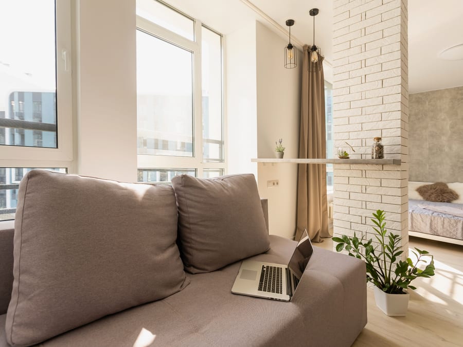 A sunlit studio apartment with a taupe modular sofa beside floor-to-ceiling windows, a white brick column with a slim counter shelf and cage pendant lights, and a bed with a sheepskin throw in the sleeping area beyond.