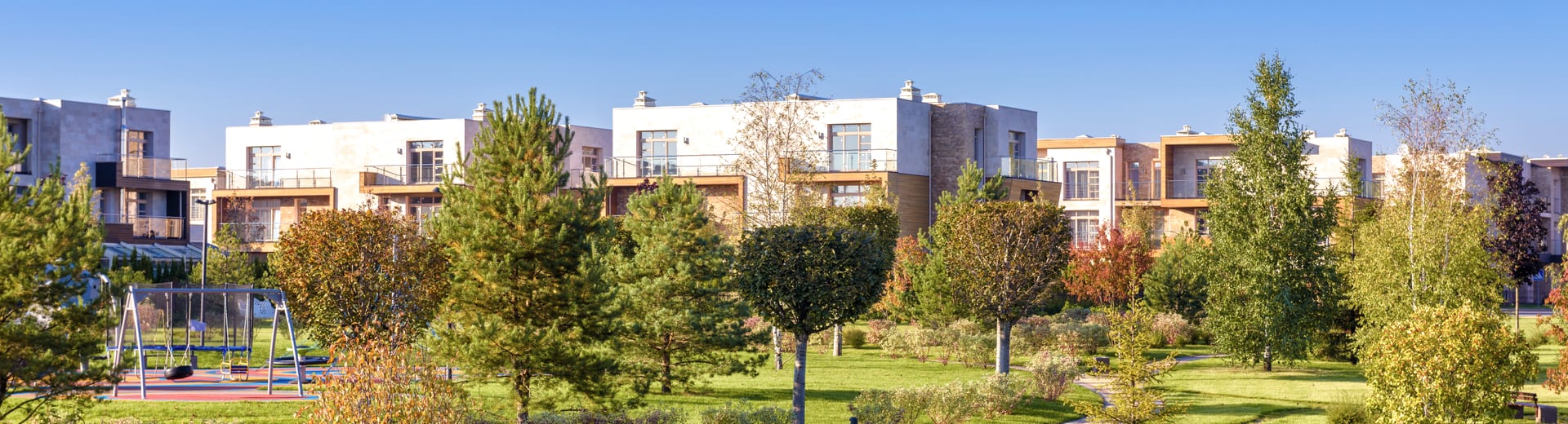 A long row of modern low-rise apartment buildings in cream stone and timber cladding, with glass-railed balconies and large windows, seen across landscaped park grounds of mown lawn, pine and birch trees, shrubs turning red and gold, and a curving paved footpath, with a playground and swing set at the left.
